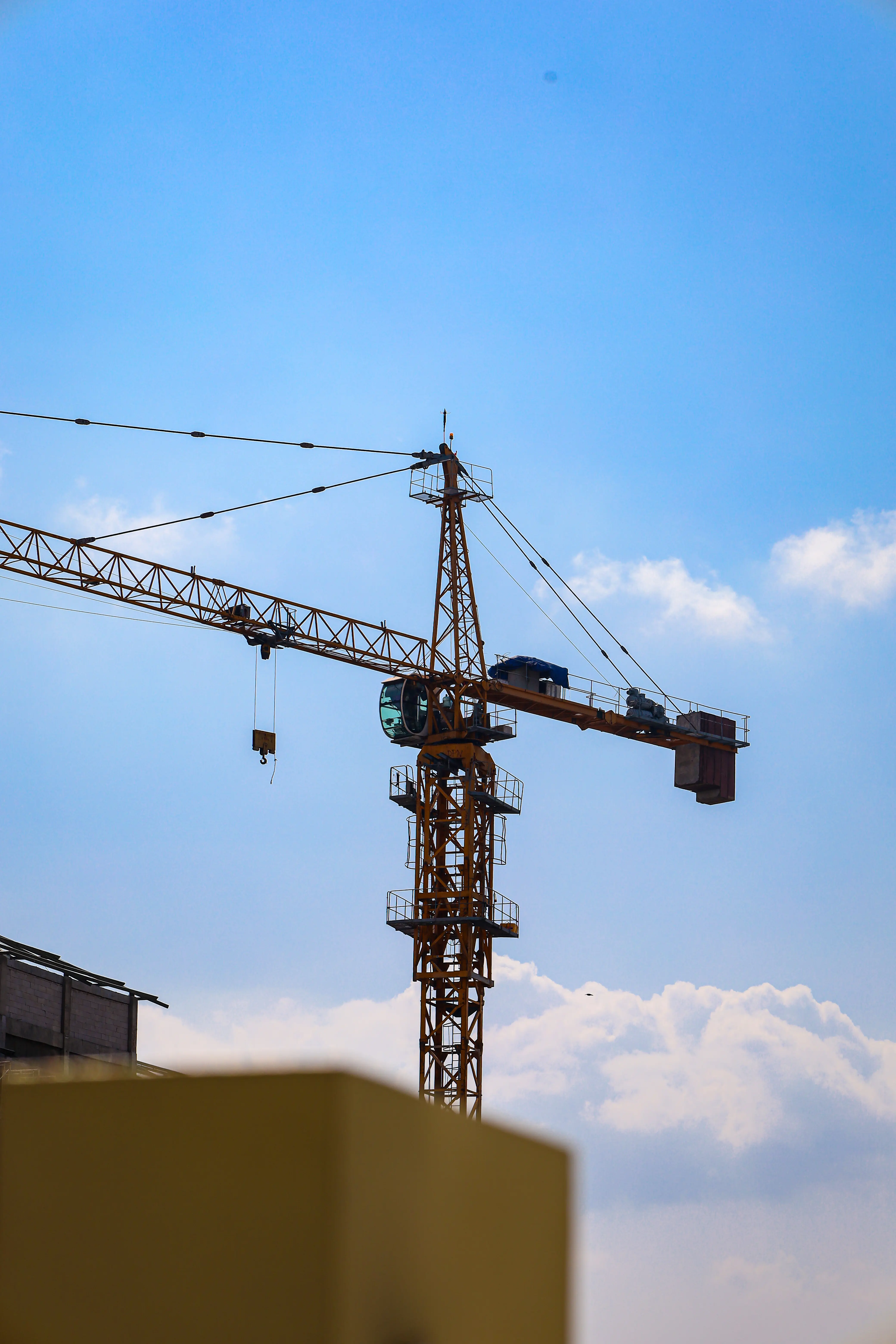 A yellow construction crane reaching across a blue sky, photographed from behind a low wall